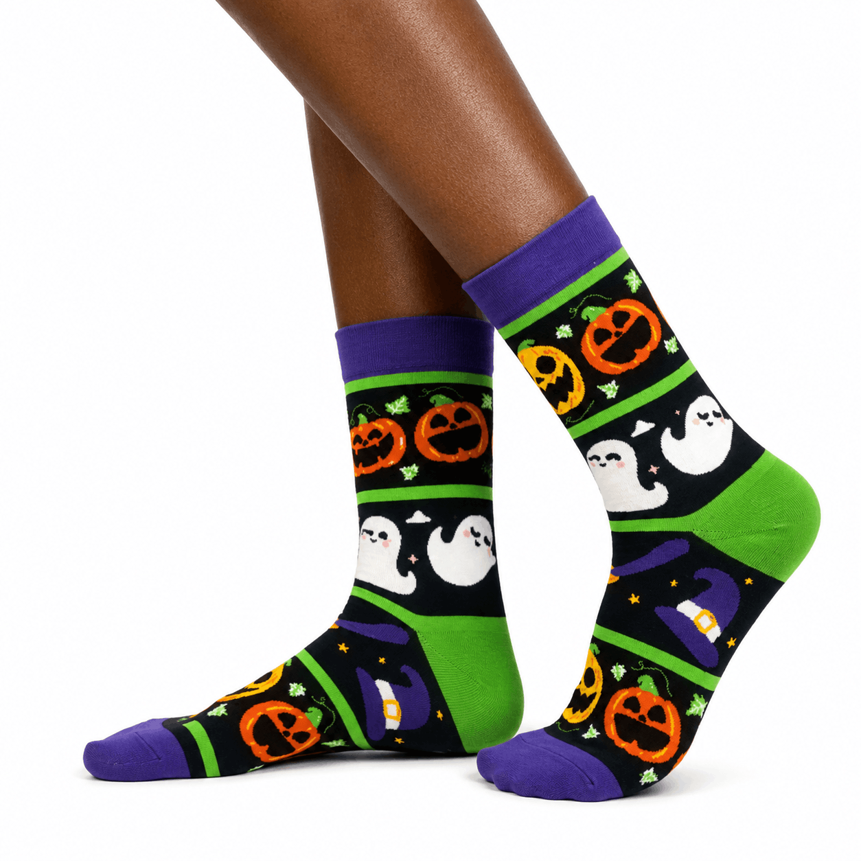 A pair of crew socks with a Halloween pattern, shown on a person's feet against a plain white background. The socks feature a black background with green stripes, orange jack-o'-lanterns, white ghosts, purple cuffs and toes, and a purple witch hat motif.